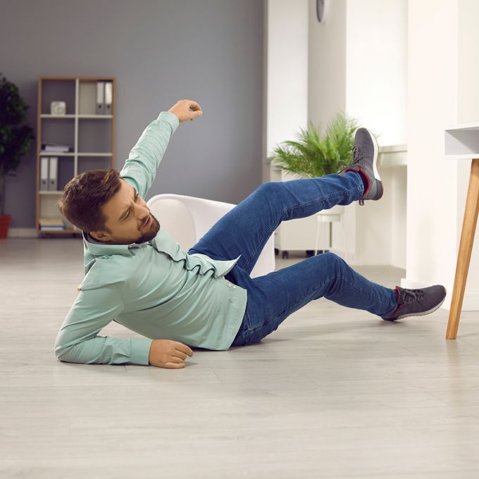 Full body photo of a young clumsy brunette man with unshaven bristles falling down to the floor and injuring his arm at home