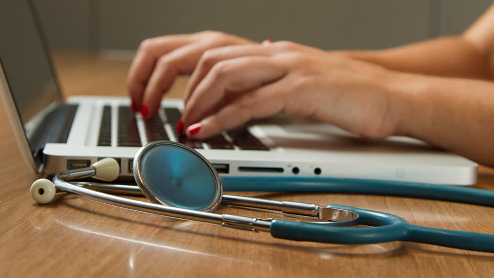 Woman typing on a computer with a stethoscope sitting next to it