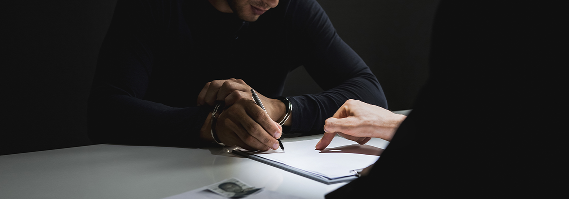 Criminal man with handcuffs signing document in interrogation room