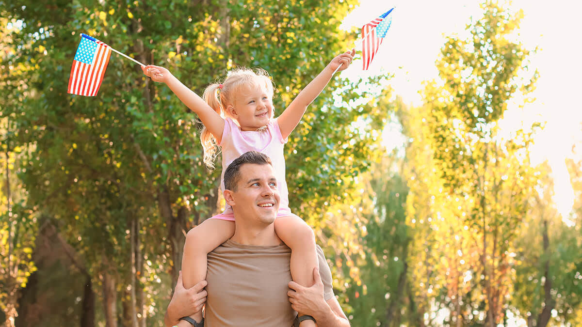 Girl sitting on her father's shoulders holding two little American flags