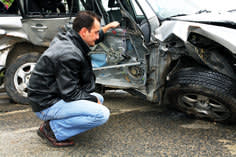 Man in a leather jacket crouched down look at his totalled car