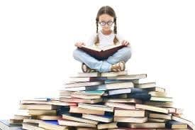 young girl with glassed and pigtails sitting on top of a giant stack of books, looking down at an open book in her lap