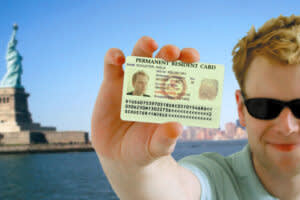 Man in sun glasses holding a card with his picture that says "permeant resident card" in front of the Statue of Liberty 