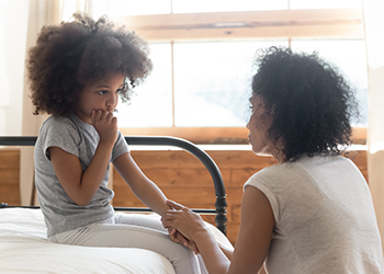 A mother holds her daughters hand while she sits on a bed