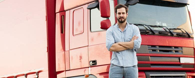 Bearded Man in button down and jeans standing in front of red semi truck with his arms crossed