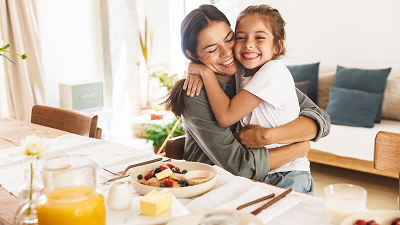 Mother and daughter smiling and hugging while eating breakfast