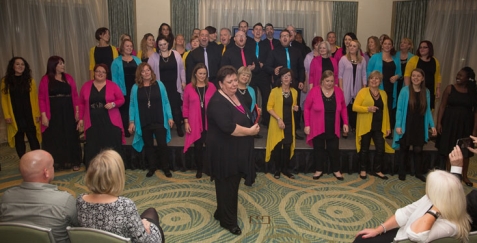 Women standing in front of a group of people in rows on steps wearing coordinated colored cardigans