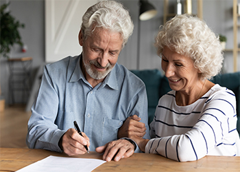 Two older people smiling while signing paperwork at desk