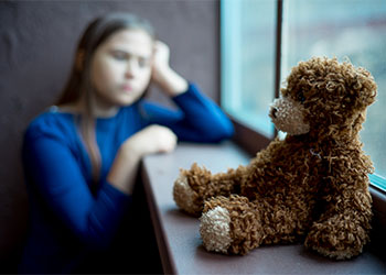 Child looking out a window with a teddy bear