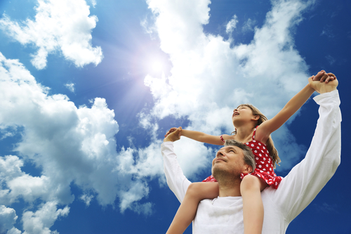 A father holding his daughter on his shoulders, looking at a cloudy sky.