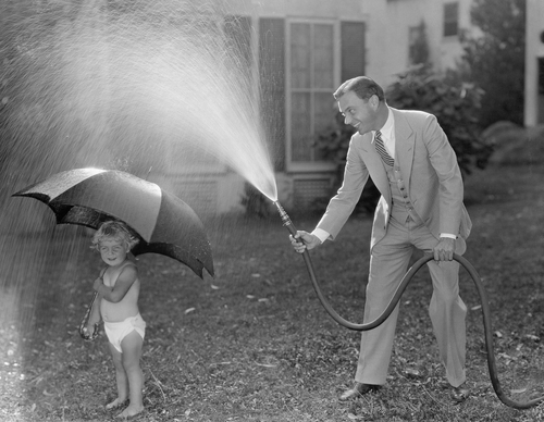 A father playing out side with his kid and a water hose.