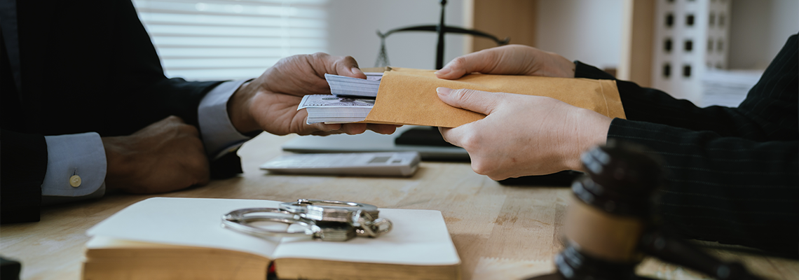 Close-up of hands exchanging a stack of cash across a desk with a gavel and handcuffs