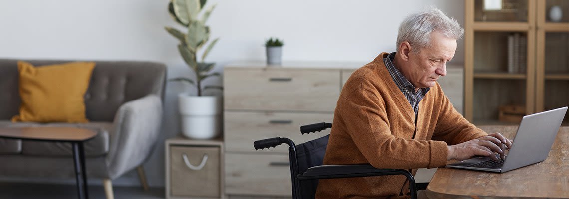 An elderly man in a wheelchair works on a laptop