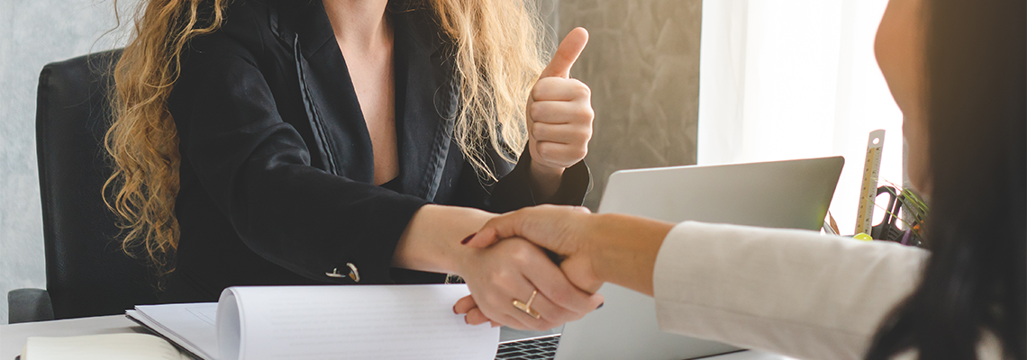 Two people sitting at desk shaking hands with thumbs up
