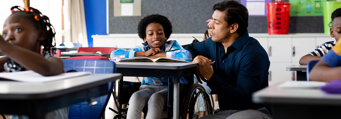 Teacher helping schoolboy sitting in wheelchair in classroom
