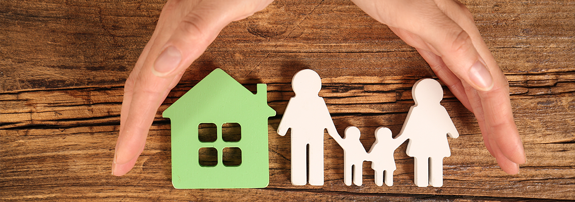 Woman holding hands near figures of house and family on wooden background