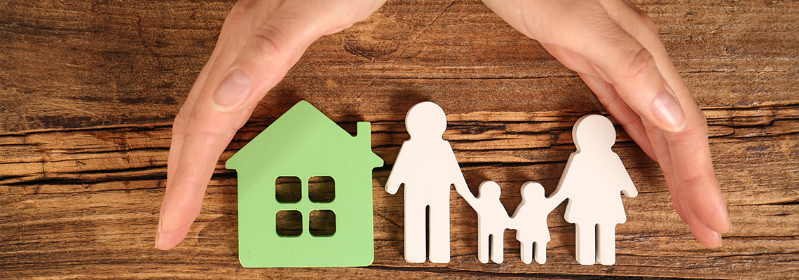 Woman holding hands near figures of house and family on wooden background