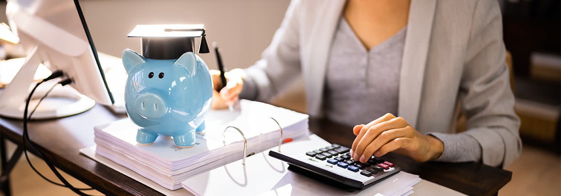 Person at desk with calculator, piggy bank and graduation cap