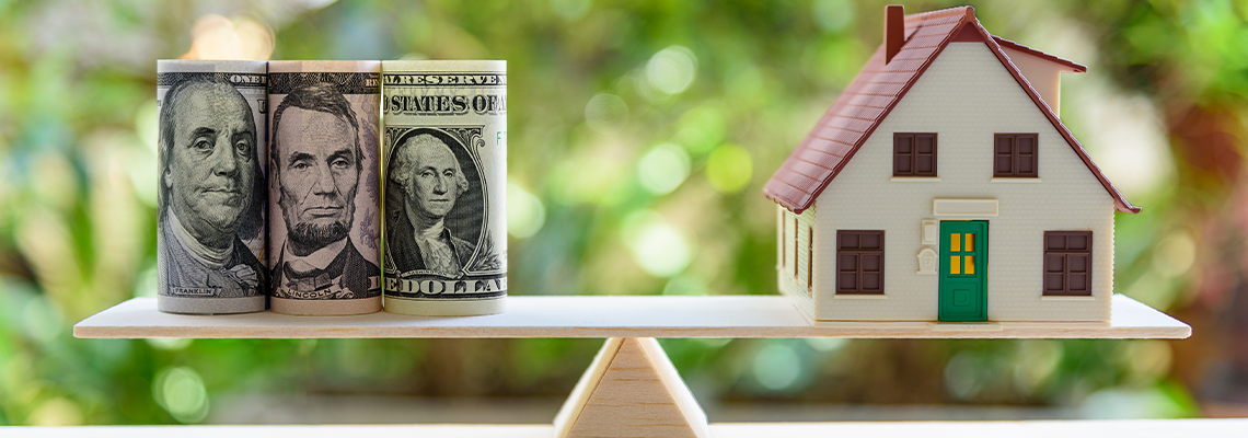 US dollar bills across from a house model on a balance scale