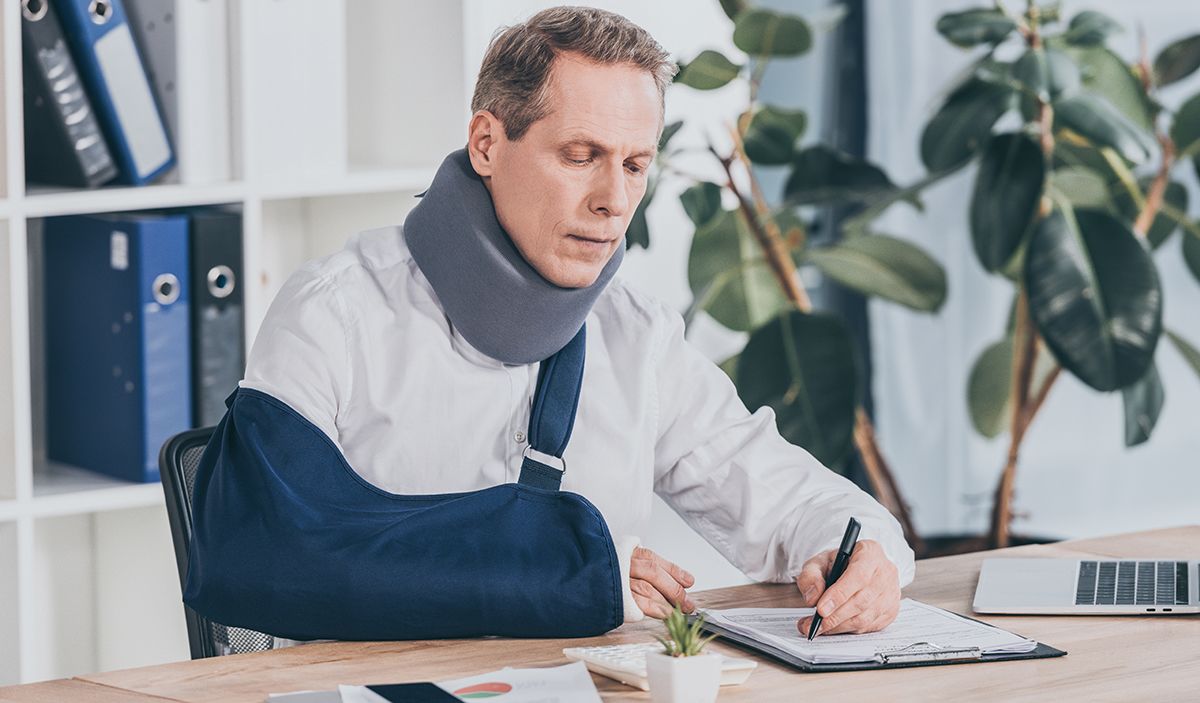 worker in neck brace and arm bandage sitting at table and carefully writing on document with pen in office