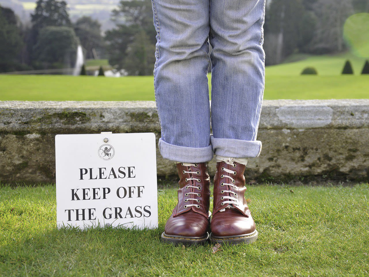Woman Stepping on The Grass with A Sign Please Step out The Grass Next to Her Feet
