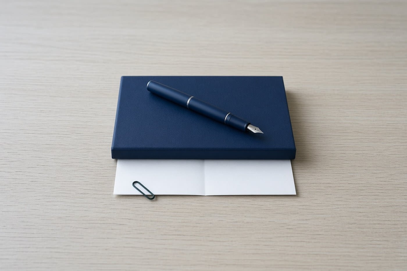 A closed navy notebook with a folded blank sheet of paper tucked partly beneath it, a navy fountain pen across the cover and a small dark green paper clip on the sheet's edge