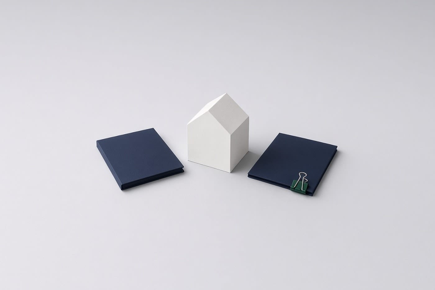 A small white block-model house standing between two closed navy folders, with a green binder clip on the right-hand folder