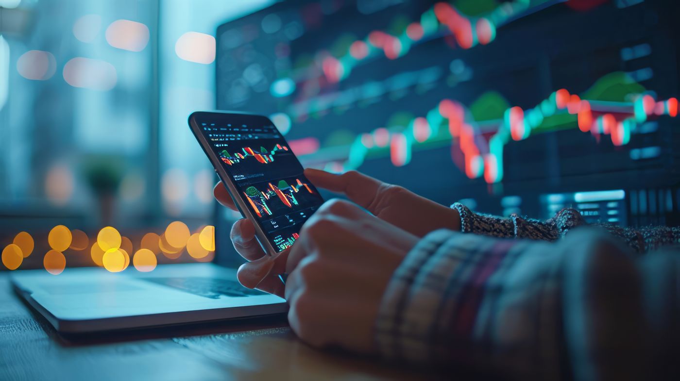 Trader hand with mobile phone and computer screen with trading candlestick
