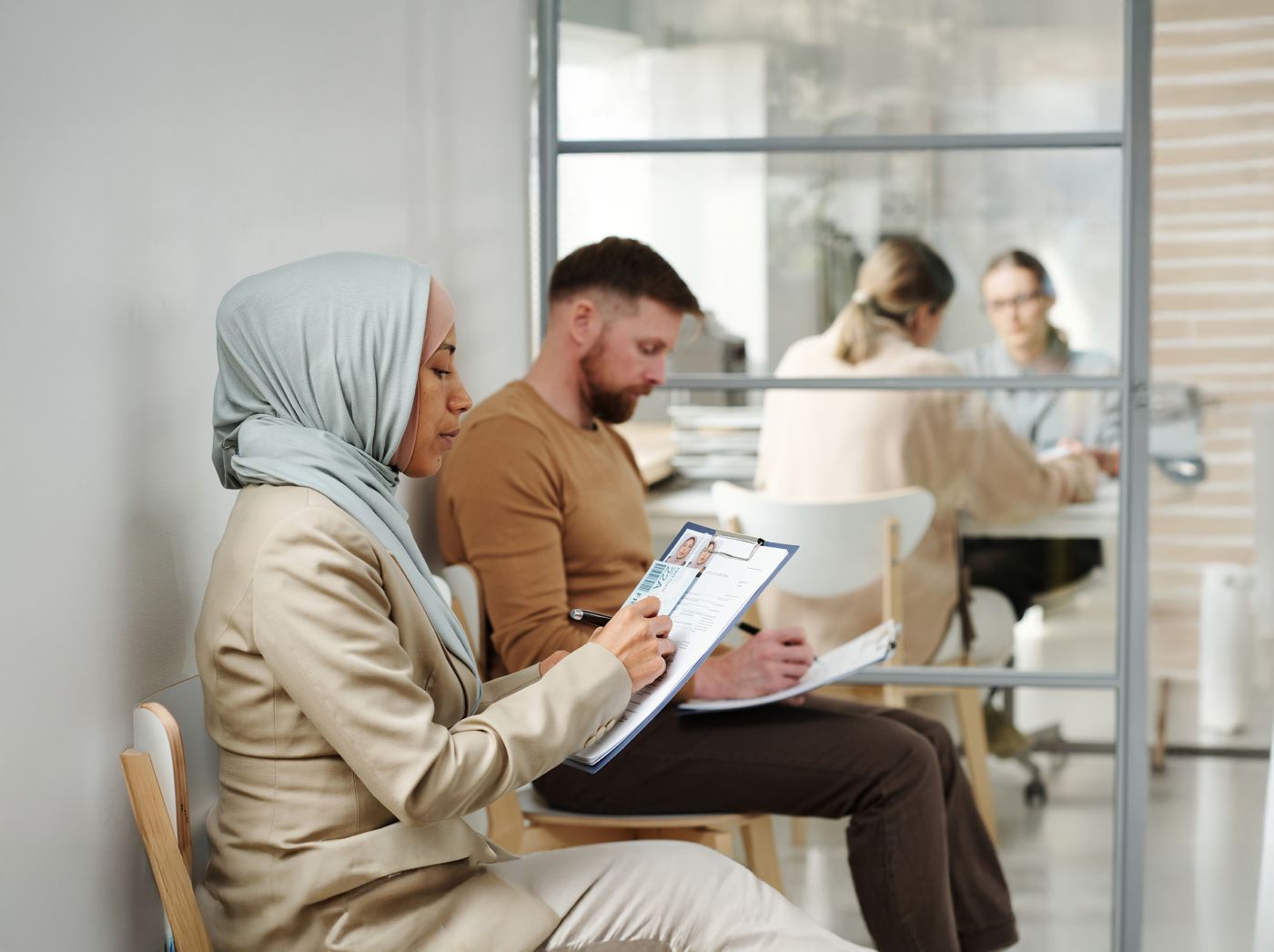 Young adult Caucasian man and Muslim woman in hijab sitting on chairs in visa service agency filling in application forms while waiting for appointment