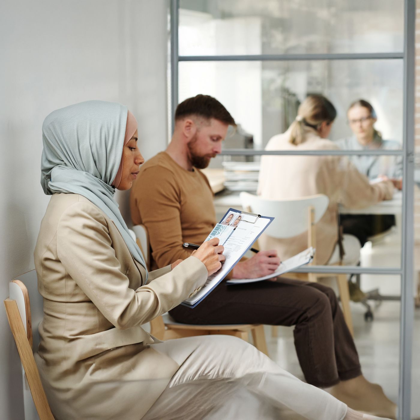 Young adult Caucasian man and Muslim woman in hijab sitting on chairs in visa service agency filling in application forms while waiting for appointment