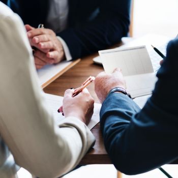 Three people at a table. One is holding a pen, one is holding paper, and one has their hands clasped. 