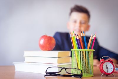 young boy sitting at desk with a stack of books and an apple on top, colored pencils, miniature clock, and glasses