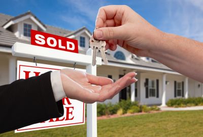 Hand dropping keys into another hand in front of sold sign outside a house