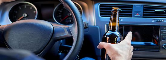 Closeup male hand is holding beer behind steering wheel of car.