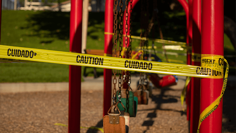 Empty saddle style swings at a playground covered in yellow caution tape written in english and spanish.