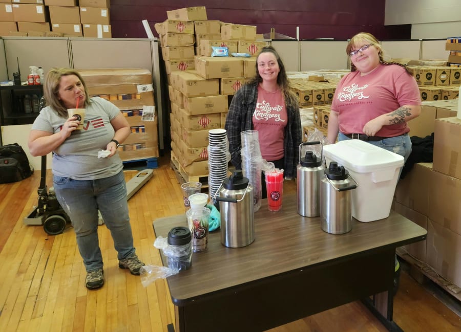Three volunteers at a small table set up with coffee thermoses, hot chocolate, and cups, surrounded by pallets of supplies in the warehouse