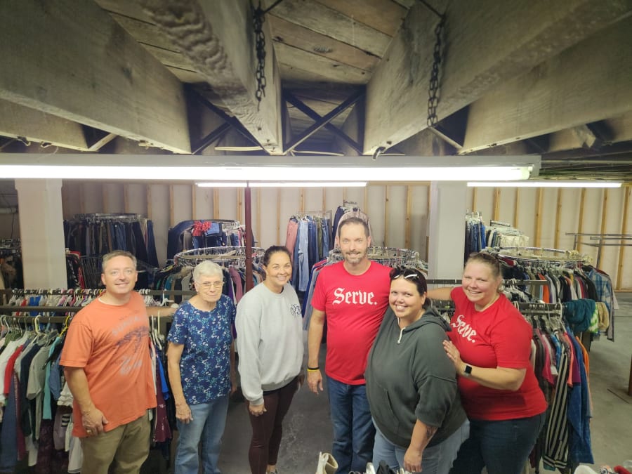 Six volunteers, several wearing matching red Serve t-shirts, posing together inside a clothing closet lined with racks of clothes