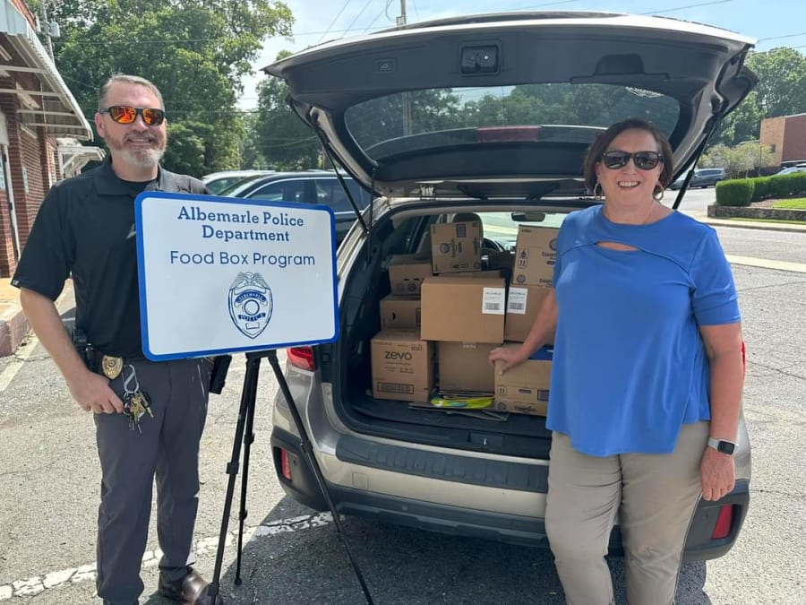 Albemarle police officer and partner standing beside the Food Box Program sign with an open SUV trunk full of supply boxes