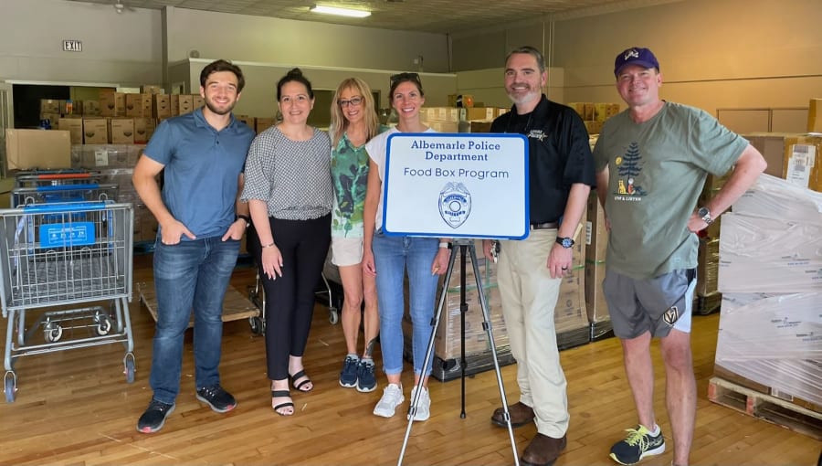 Group of volunteers and officers gathered around the Albemarle Police Department Food Box Program sign inside the Badges & Boxes warehouse
