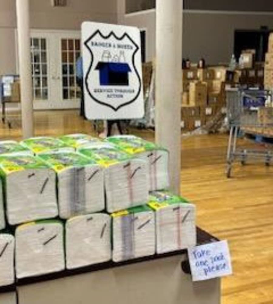 The Badges & Boxes 'Service Through Action' sign on a stand next to stacks of paper-towel packs with a handwritten note reading Take One Pack, Please