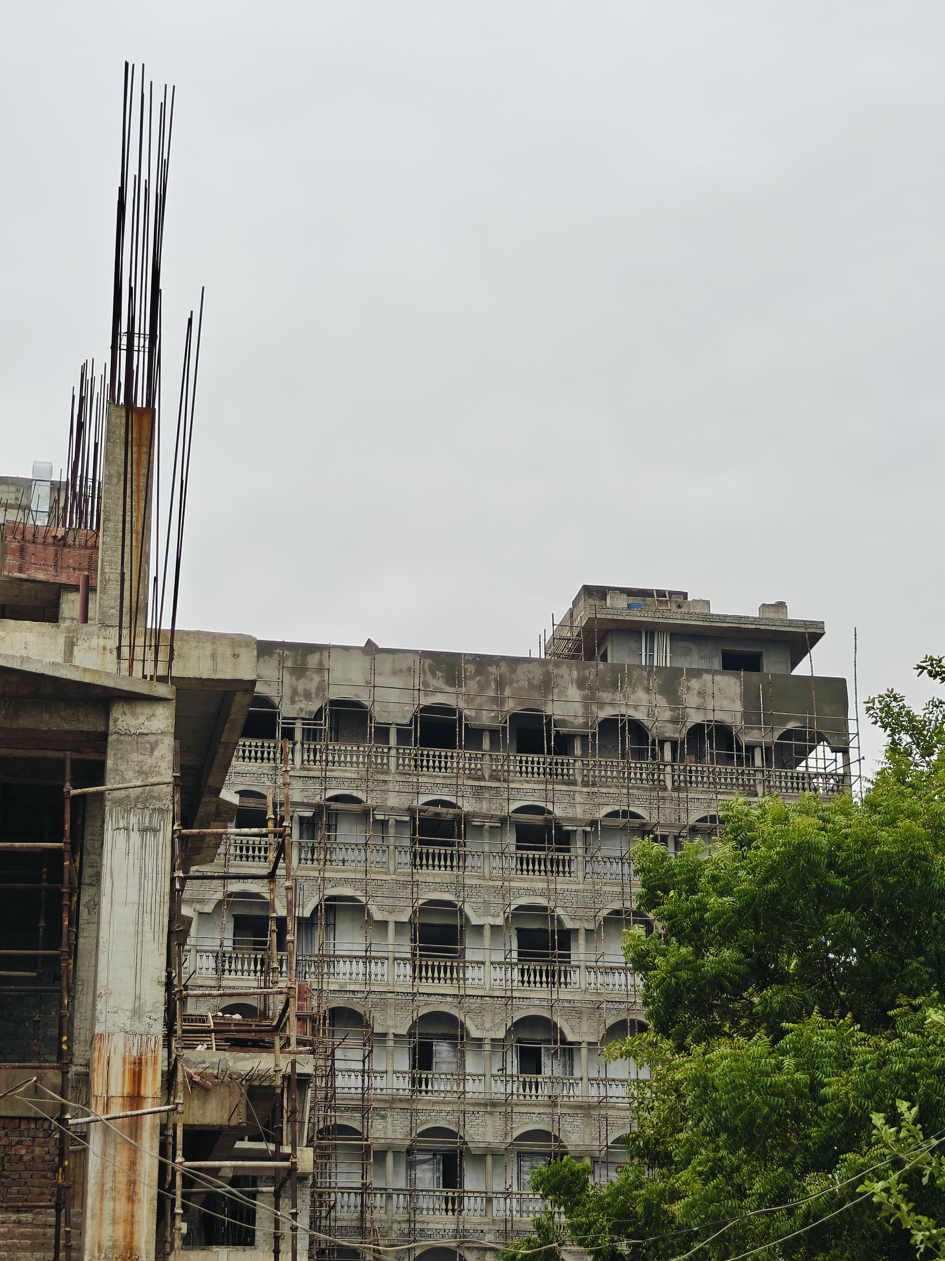 Close-up of an unfinished concrete column with exposed rebar at the ISKCON Wave City site, with a scaffolded building with arched balconies in the background