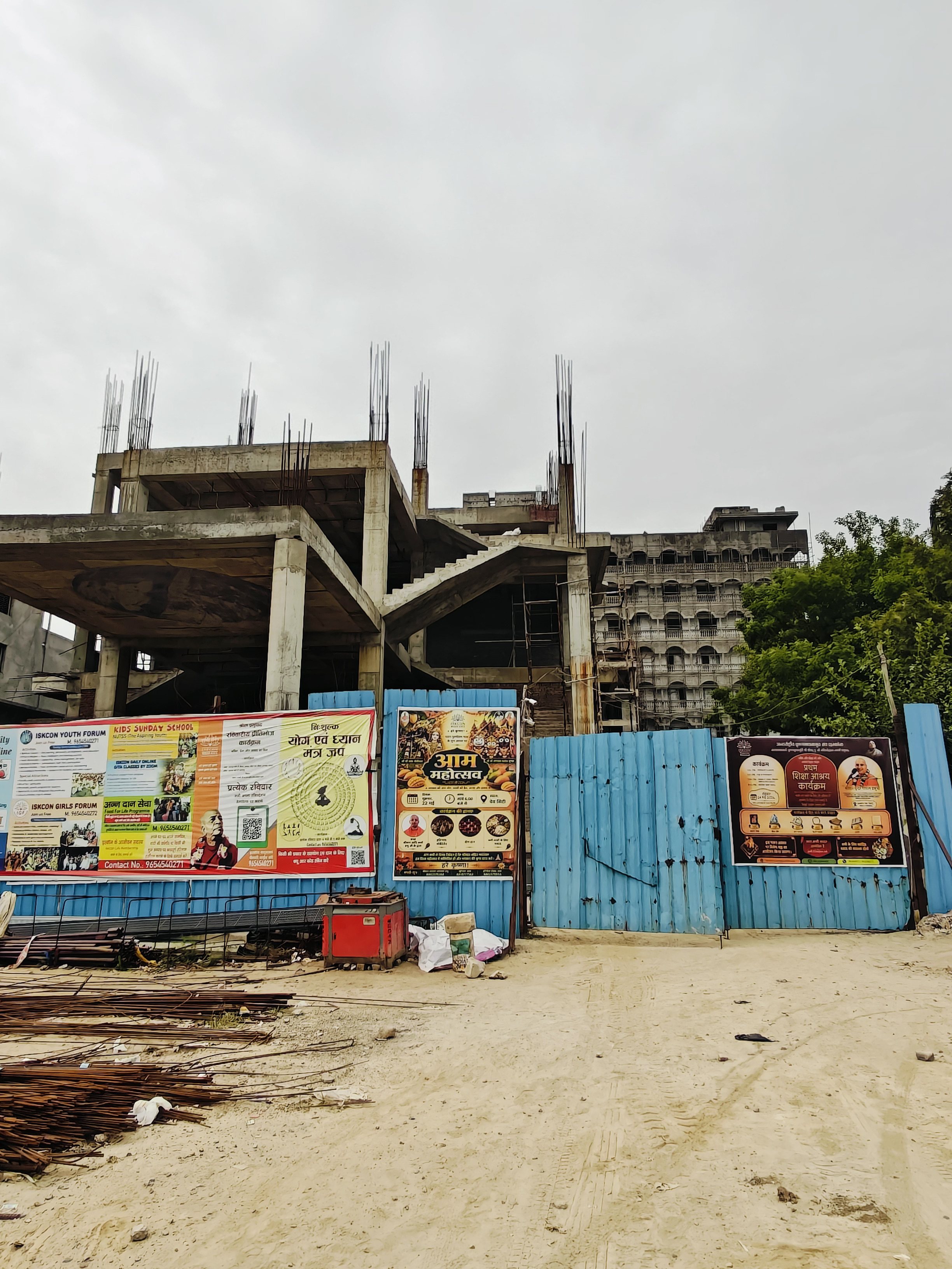 ISKCON Temple Wave City site entrance with blue tin fencing covered in event banners — ISKCON Youth Forum, Kids Sunday School, Yog evam Dhyan Mantra Jaap, and Aam Mahotsav — with the under-construction temple structure behind