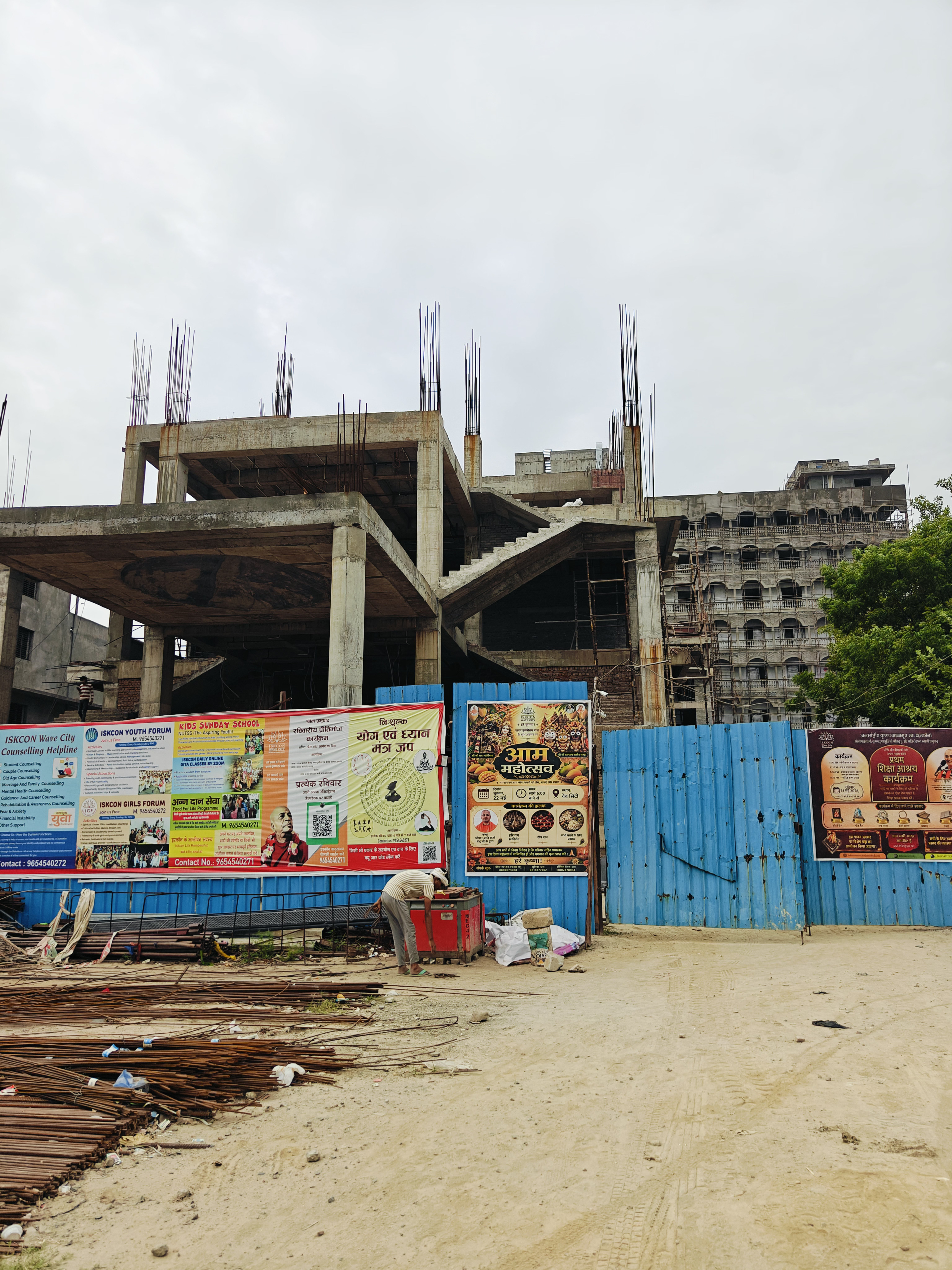 ISKCON Temple Wave City under construction — exposed rebar columns and unfinished concrete staircase, with the completed multi-storey building visible behind blue site fencing