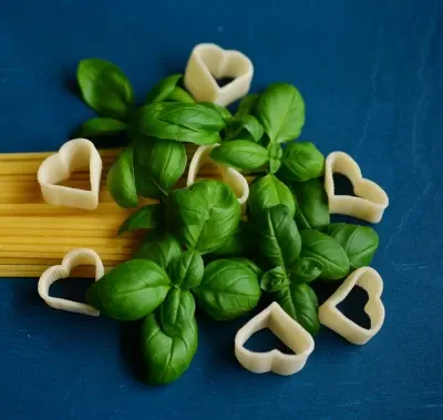 Basil leaves and heart shaped pasta pieces on a blue background.