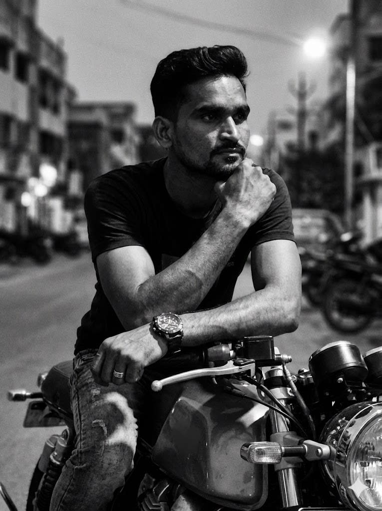 A black-and-white portrait with a rugged, cinematic aesthetic captures a male figure seated on a classic motorcycle (café racer or bobber style). The subject is leaning forward over the handlebars, striking a thoughtful and intense pose. One hand rests on the headlight or handlebars, while the other arm is bent, supporting his head. His gaze is directed outward, conveying a contemplative and virile expression.
He wears a dark cotton T-shirt and pants (possibly jeans). A chunky wristwatch is visible on one arm, and a ring on his finger.
His hair is medium-length, textured, and casually tousled. A short, well-groomed beard defines his face.
The background is a softly blurred urban or street environment. The lighting is natural and dramatic, with high contrast, creating deep shadows and sharp highlights that sculpt the facial features and arm muscles. The image has a subtle grain, evoking the look of analog film.
Camera Settings: Captured with a prime portrait lens (e.g., 85mm or 105mm) on a full-frame camer