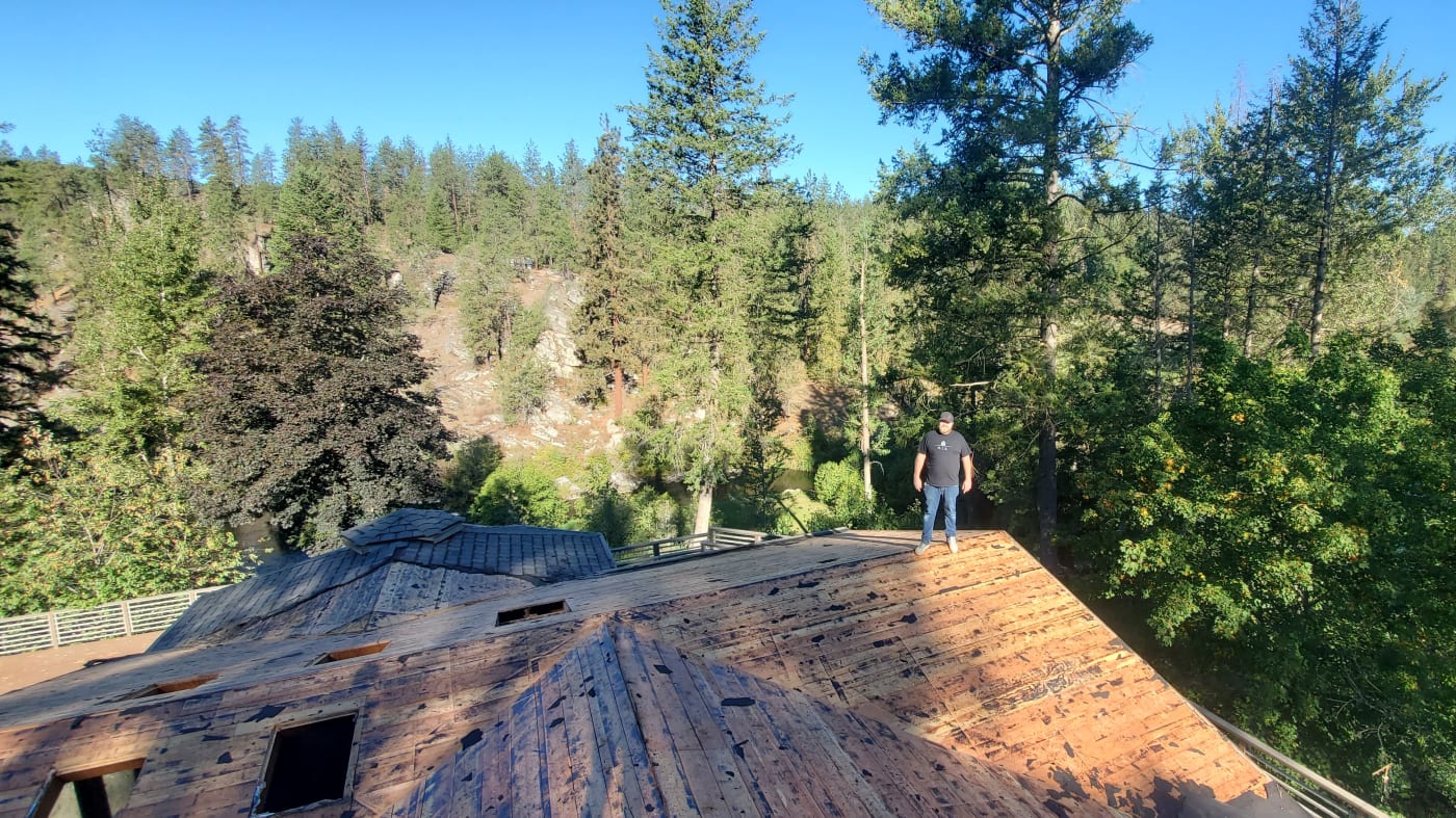 Nathan Taylor, the founder and owner of NIB, standing confidently on the roof of a custom home in the Spokane Metro WA area. Surrounded by a scenic backdrop of pine trees, he ensures that the high standards NIB is known for are upheld, emphasizing commitment to efficiency and budget management on every project.