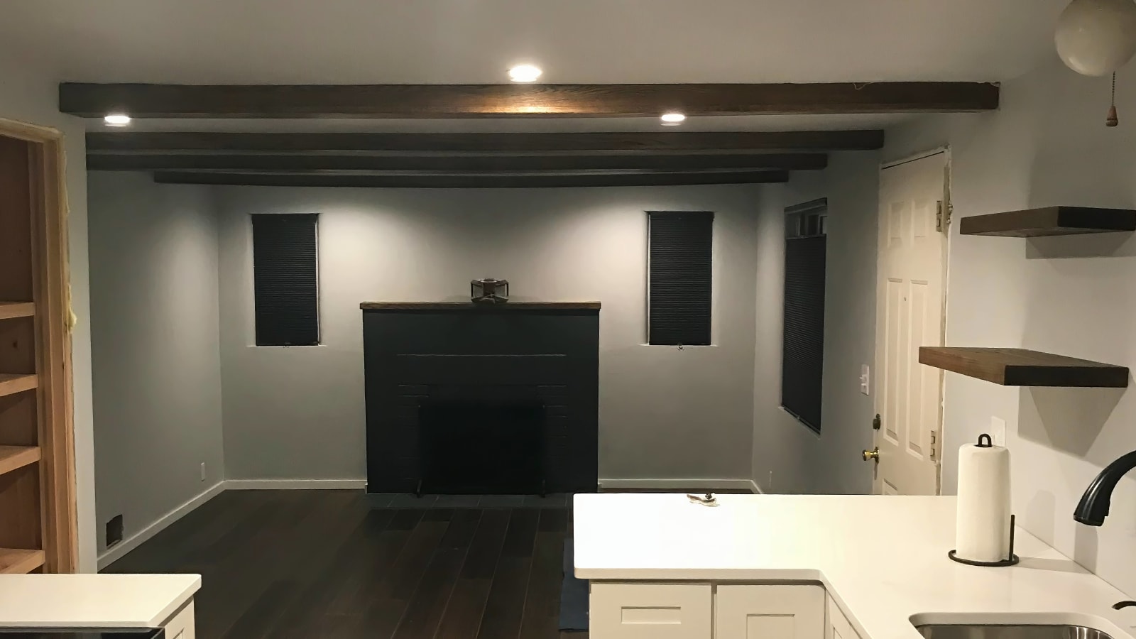 Cozy modern living room done by NIB in the Spokane, WA area, features custom wood beams that add warmth and character to the space. The sleek black fireplace creates a striking contrast with the light gray walls, while the recessed lighting brings out the elegance of the room. Dark wood flooring ties everything together, creating a sophisticated, yet inviting atmosphere. The subtle floating shelves offer a contemporary touch, perfect for displaying personal décor, all contributing to a thoughtfully remodeled space.