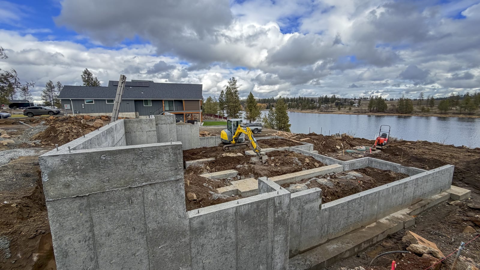 An NIB custom home project in Spokane, WA, during the foundational phase. The foundation walls are constructed from poured concrete, and an excavator is actively working on the site, preparing the ground for the next steps. In the background, the nearby waterfront adds a scenic touch, with trees and another home visible. The construction site is still in the process of leveling and preparing the land. This shows the crucial early stages of home building, with careful attention to the site layout and preparation by NIB’s skilled team.