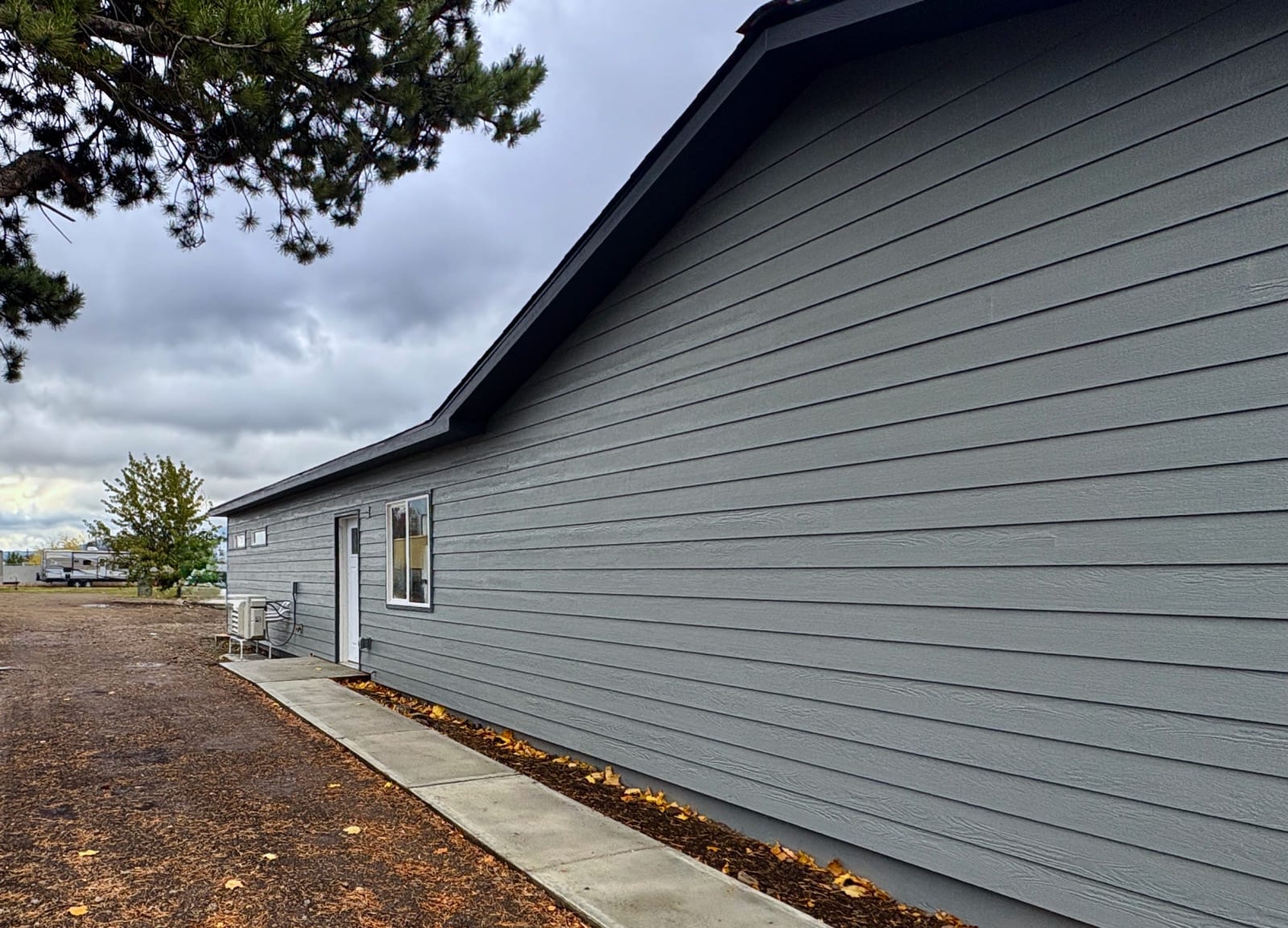Finished exterior of a newly built home showcasing skilled craftsmanship, with crisp gray horizontal lap siding running the length of the structure and a clean dark roofline overhead. A white door and window are neatly trimmed against the siding, with a poured concrete walkway leading to the entrance. A pine tree frames the top left of the shot, with a mobile home and cloudy sky visible in the background — evidence of quality construction and attention to detail from foundation to finish.