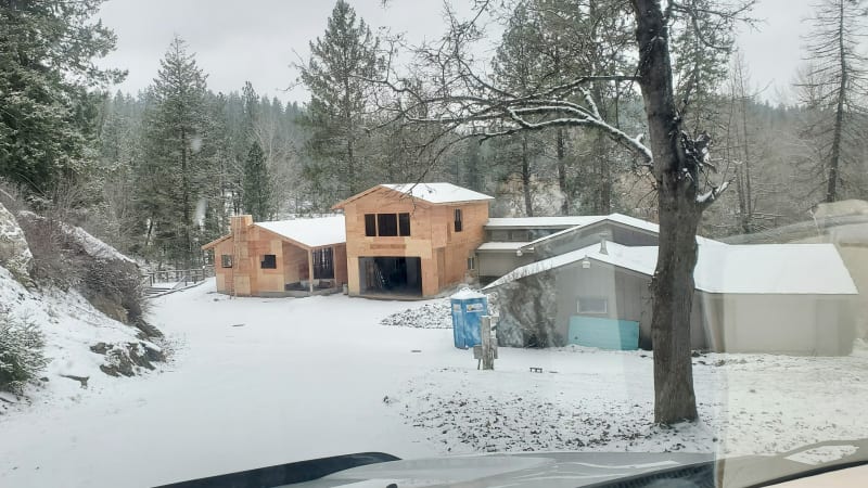 A winter scene showing the progress of a custom home addition in the Spokane Metro WA area, done by NIB. The addition is framed but still awaiting sheathing, with open window spaces and rooflines exposed. The snow-covered ground and surrounding trees add a serene, seasonal backdrop to the active construction site.